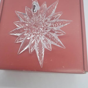 May include: Clear glass star-shaped ornament with intricate detailing. The ornament has pointed edges and a central circular design. It hangs from a red ribbon and is placed on a red box.