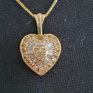 Puede incluir: Un collar con colgante en forma de coraz&oacute;n de color dorado. El colgante presenta un dise&ntilde;o en forma de coraz&oacute;n con un coraz&oacute;n m&aacute;s peque&ntilde;o en el centro, rodeado de piedras transparentes y un patr&oacute;n de rayos de sol. La cadena del collar tambi&eacute;n es dorada.