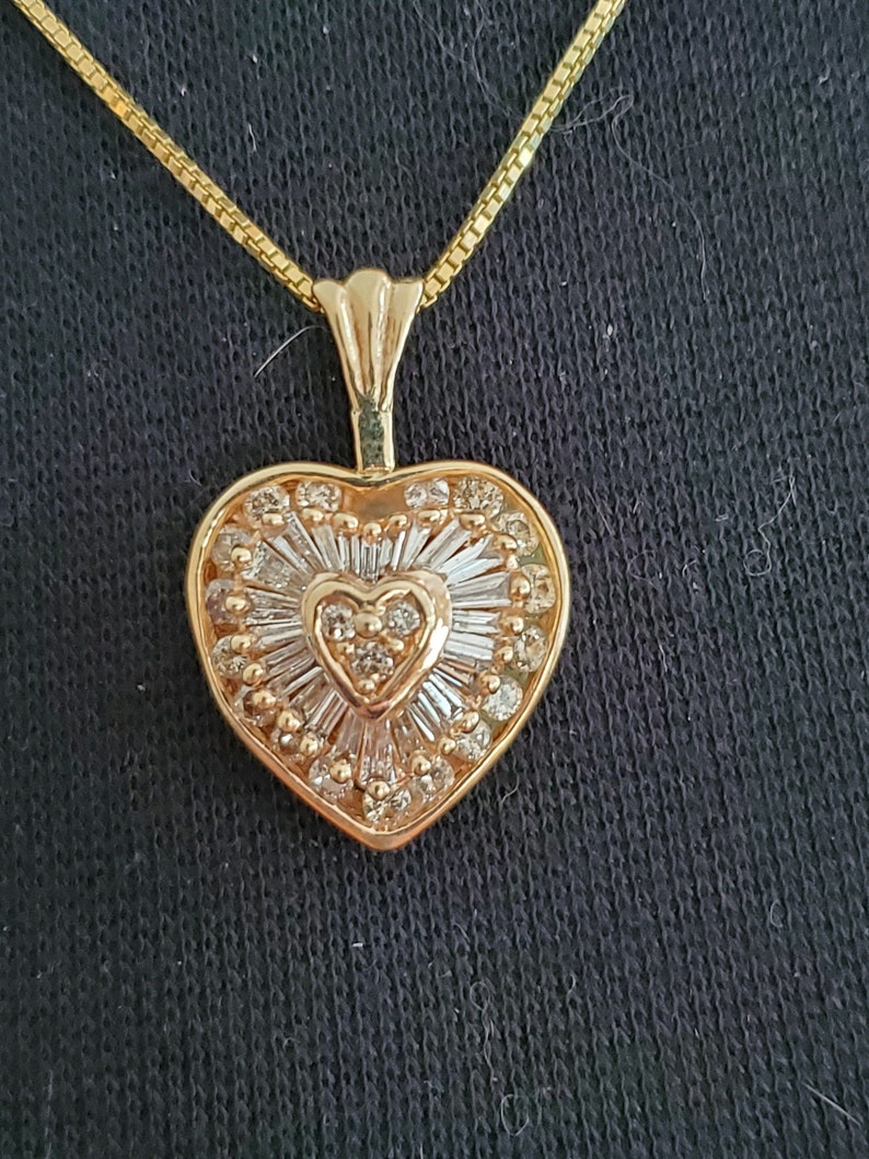Puede incluir: Un collar con colgante en forma de coraz&oacute;n de color dorado. El colgante presenta un dise&ntilde;o en forma de coraz&oacute;n con un coraz&oacute;n m&aacute;s peque&ntilde;o en el centro, rodeado de piedras transparentes y un patr&oacute;n de rayos de sol. La cadena del collar tambi&eacute;n es dorada.