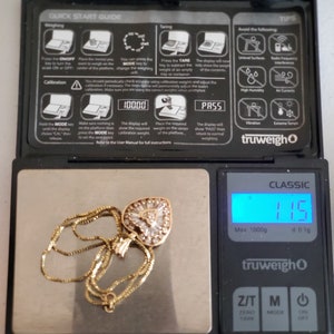 Puede incluir: Un collar de oro con un colgante en forma de coraz&oacute;n, engastado con piedras transparentes, descansa sobre una balanza digital. La balanza muestra un peso de 11,5, con una capacidad m&aacute;xima de 1000g. La balanza tiene un cuerpo negro y una pantalla digital.