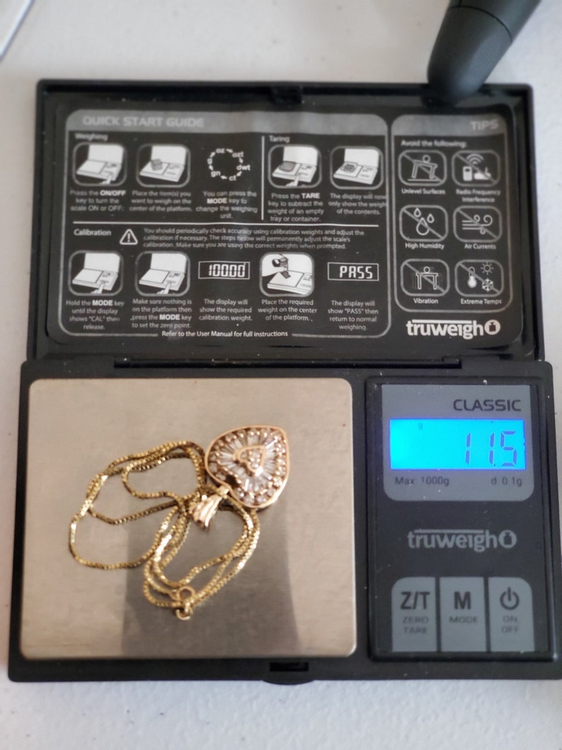 Puede incluir: Un collar de oro con un colgante en forma de coraz&oacute;n, engastado con piedras transparentes, descansa sobre una balanza digital. La balanza muestra un peso de 11,5, con una capacidad m&aacute;xima de 1000g. La balanza tiene un cuerpo negro y una pantalla digital.