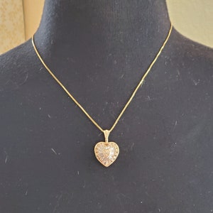 Puede incluir: Un collar dorado con un colgante en forma de coraz&oacute;n. El colgante est&aacute; adornado con peque&ntilde;as piedras brillantes y tiene un dise&ntilde;o detallado. El collar se exhibe en un maniqu&iacute; negro.