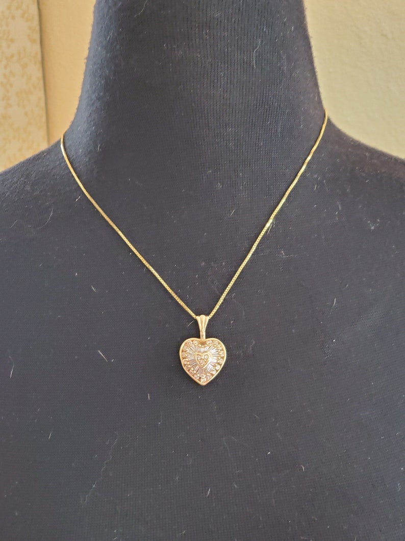 Puede incluir: Un collar dorado con un colgante en forma de coraz&oacute;n. El colgante est&aacute; adornado con peque&ntilde;as piedras brillantes y tiene un dise&ntilde;o detallado. El collar se exhibe en un maniqu&iacute; negro.