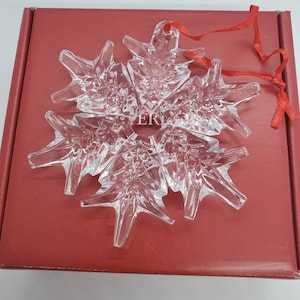 May include: A clear, crystal snowflake ornament with six points, a red ribbon, and the letters "ER" etched in the center. The ornament is resting on a red box.