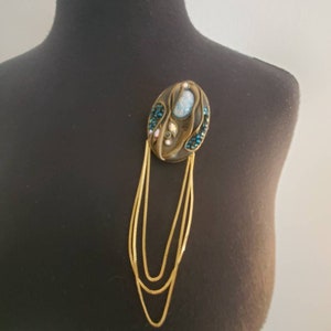 May include: An oval-shaped brooch with a bronze-colored base, adorned with blue and pink gemstones and a central light blue stone. Three strands of gold-colored chain hang from the brooch, creating a layered effect. The brooch is attached to a black mannequin.