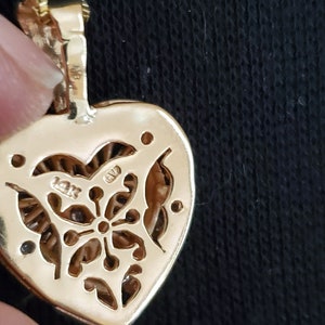 Puede incluir: Primer plano de un colgante de oro en forma de coraz&oacute;n con un dise&ntilde;o floral en la parte posterior. El colgante est&aacute; sujeto a una cadena de oro. El colgante est&aacute; marcado "14K".