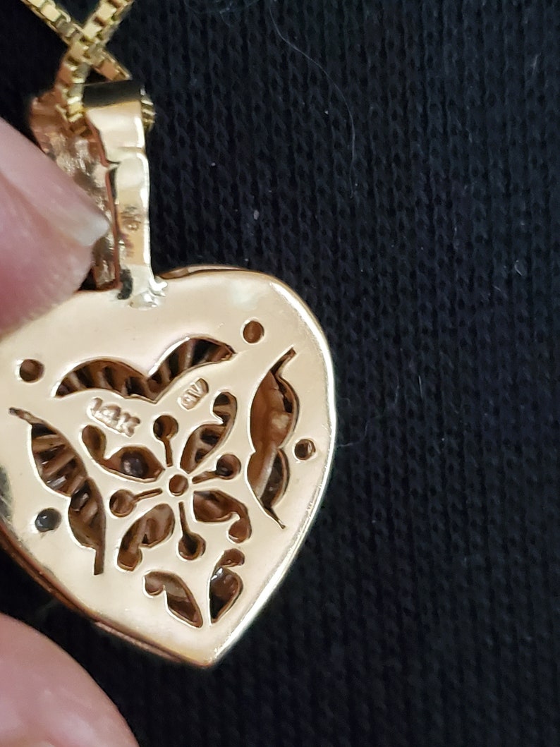 Puede incluir: Primer plano de un colgante de oro en forma de coraz&oacute;n con un dise&ntilde;o floral en la parte posterior. El colgante est&aacute; sujeto a una cadena de oro. El colgante est&aacute; marcado "14K".