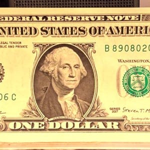 May include: A United States one dollar bill featuring a portrait of George Washington in the center, the text THE UNITED STATES OF AMERICA, and serial number B 89080206 C. A small orange square sticker with a black arrow is attached to the right side.