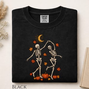 May include: A black t-shirt featuring a graphic of two skeletons dancing under a yellow crescent moon. The design includes scattered orange autumn leaves and three orange pumpkins at the bottom. The inner neck label reads Comfort Colors.