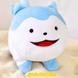 May include: A plush toy shaped like a creature with light blue and white fur, featuring two black eyes, a small black nose, and a wide open mouth with a red interior. The text @MioStitches is printed in yellow at the bottom of the plush toy.