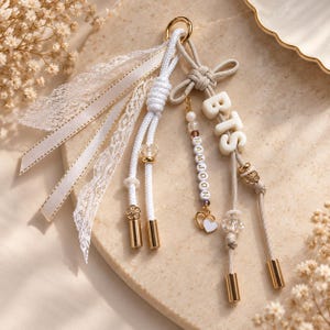 May include: A decorative keychain featuring white and beige cords, lace ribbons, and gold-toned metal accents. It includes white letter beads spelling BTS and NAMJOON, clear crystal-like beads, a small heart charm, and gold-toned metal end caps.