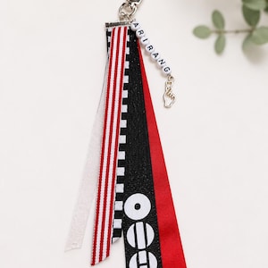 May include: A ribbon keychain featuring a silver lobster clasp, a string of white beads spelling ARIRANG, and a silver finger heart charm. The accessory includes multiple fabric ribbons in white, red and white stripes, black and white checks, and solid red.