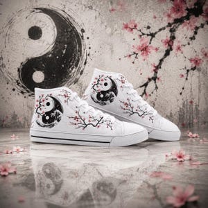 Könnte beinhalten: Ein Paar weiße High-Top-Sneaker mit einer schwarz-weißen Yin-Yang-Grafik. Das Design zeigt zarte rosa Kirschblütenzweige, die sich über das Obermaterial aus Segeltuch erstrecken, ergänzt durch weiße Schnürsenkel und eine weiße Gummisohle.