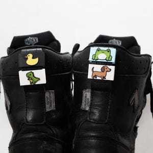Könnte beinhalten: Ein Paar schwarze Stiefel mit hohem Schaft und dekorativen rechteckigen Anhängern an den hinteren Zuglaschen. Die Anhänger zeigen farbenfrohe Illustrationen einer gelben Ente, eines grünen Dinosauriers, eines Frosches und eines braunen Dackels.
