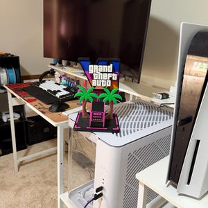 May include: A 3D printed Grand Theft Auto V logo sign featuring a black base, two green palm trees, and a pink wanted level display with five stars. The sign has a blue, purple, and black gradient background with white text reading Grand Theft Auto.