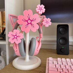 Op de afbeelding: Een witte hoofdtelefoonstandaard met een kattenoordesign, versierd met drie roze kersenbloesems. De standaard houdt een roze en grijze hoofdtelefoon vast, waarbij de bloemen aan de bovenkant en zijkanten van het kattenoorframe zijn geplaatst.