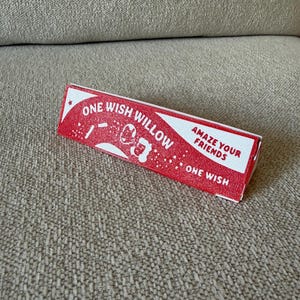 May include: A rectangular red and white paper box with a textured surface. The text reads ONE WISH WILLOW, AMAZE YOUR FRIENDS, and ONE WISH. The design includes white graphic elements of stars and a stylized figure on the front of the packaging.