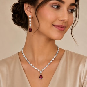 Op de afbeelding: Een zilverkleurige ketting en bijpassende oorbellen met heldere, marquise-geslepen stenen in een ketting. De ketting en oorbellen eindigen elk in een enkele, dieprode, peervormige steen die aan een heldere, peervormige steen is bevestigd.