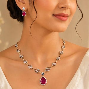 Op de afbeelding: Een gouden ketting en bijpassende oorbellen met druppelvormige rode edelstenen omringd door kleine witte steentjes. De ketting bestaat uit herhalende gebogen gouden schakels die zijn geaccentueerd met witte steentjes en een centrale hanger.