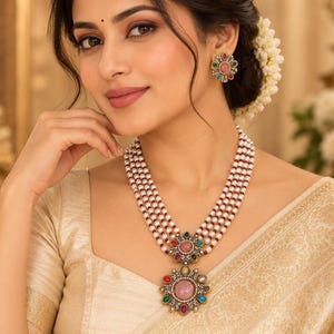 May include: A multi-strand necklace featuring rows of small white pearls and tiny red beads, centered with a large floral-shaped pendant containing a central pink stone surrounded by colorful gems. Matching floral-shaped stud earrings are included.