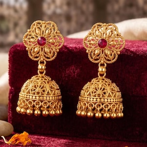 May include: A pair of gold-toned jhumka earrings featuring an intricate filigree bell-shaped base with dangling beads and a floral-shaped stud top centered with a single round red stone. The earrings exhibit a detailed metalwork pattern throughout.