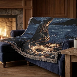 May include: A woven throw blanket featuring a dark blue and black scene of a hooded figure fishing by a campfire at night. The blanket has a fringed edge and is draped over a dark blue sofa. The design depicts a person sitting near a bright fire.