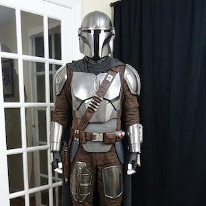 Peut inclure: Un costume complet de Mandalorien comprenant un casque, une plaque de poitrine, des &eacute;paulettes et des gantelets en argent m&eacute;tallique. La tenue se compose d'un pantalon et d'une chemise &agrave; manches longues marron, d'une ceinture utilitaire et de bottes.