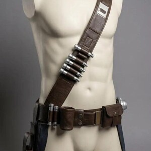 May include: A brown leather utility belt and shoulder strap set featuring silver-colored cylindrical canisters attached to the chest band. The gear includes a waist belt with multiple pouches, a holster, and a metallic buckle, worn on a mannequin.