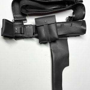 May include: A black leather utility belt featuring a series of loops for ammunition, a rectangular pouch with a snap closure, and a long holster attachment. The belt includes a metal buckle and reinforced stitching throughout the construction.