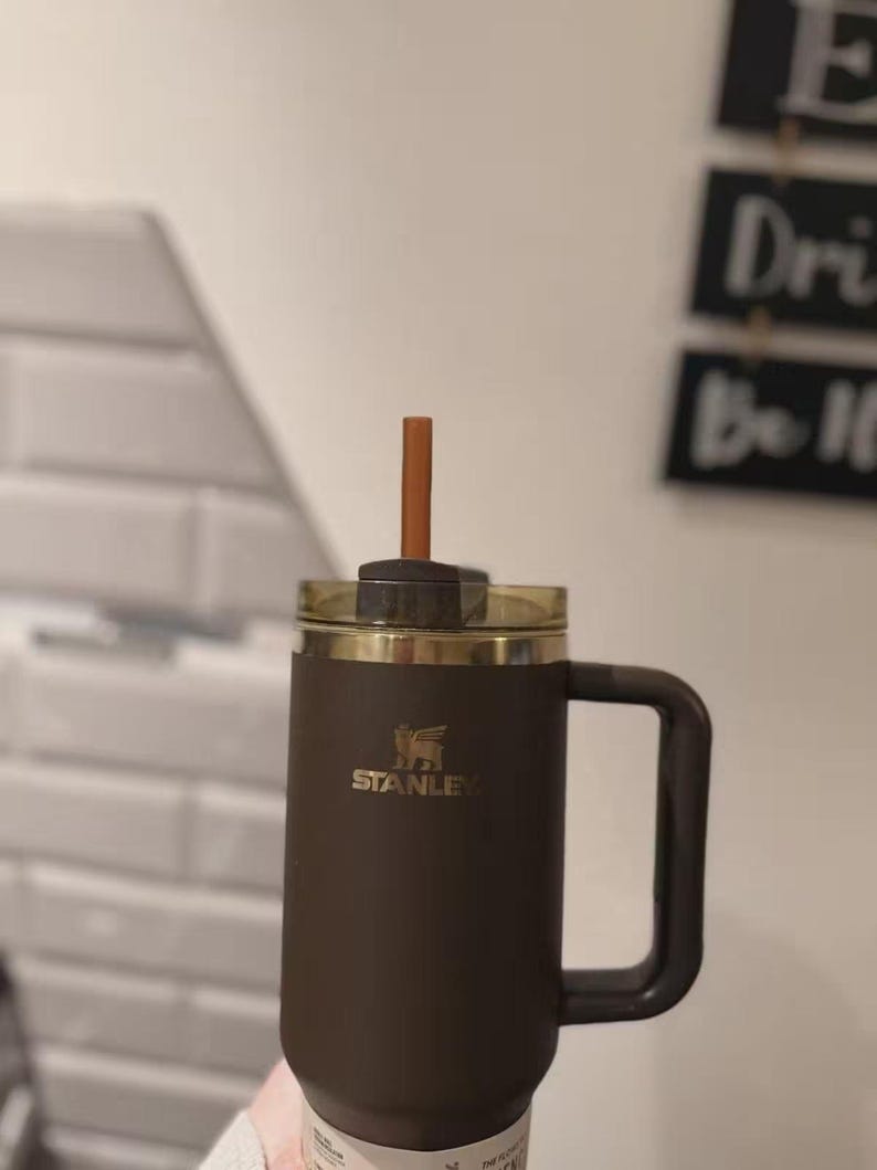 May include: A dark brown Stanley tumbler with a matching handle, a clear lid, and a brown straw. The Stanley logo is printed in gold on the front of the cup. The tumbler has a cylindrical body and a sturdy side handle for holding the beverage container.