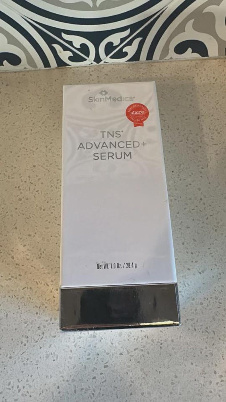 May include: A rectangular white SkinMedica TNS Advanced+ Serum box with a silver metallic base. The front features the brand logo, product name, and a red Allure Best of Beauty Hall of Fame seal. The net weight is clearly printed as 1.0 oz. / 28.4 g.