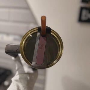 May include: A top-down view of a dark brown tumbler lid featuring a translucent olive-green rim, a dark brown handle, and a central sliding closure mechanism in a muted plum color. A thin, translucent amber-colored straw protrudes from the center.