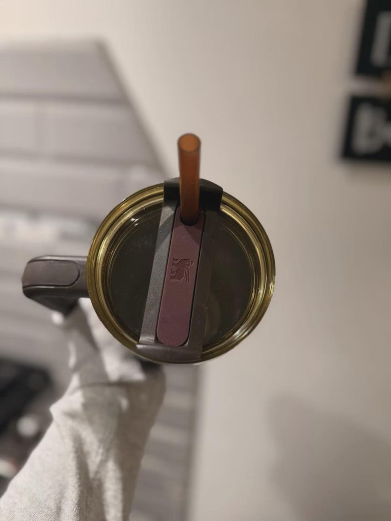May include: A top-down view of a dark brown tumbler lid featuring a translucent olive-green rim, a dark brown handle, and a central sliding closure mechanism in a muted plum color. A thin, translucent amber-colored straw protrudes from the center.