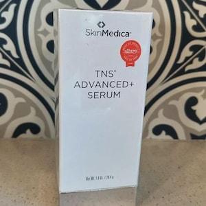 May include: A white rectangular SkinMedica TNS Advanced+ Serum box with a silver base. The front features the brand logo, product name, and a red Allure Best of Beauty seal. The text at the bottom indicates a net weight of 1.0 oz. / 28.4 g.