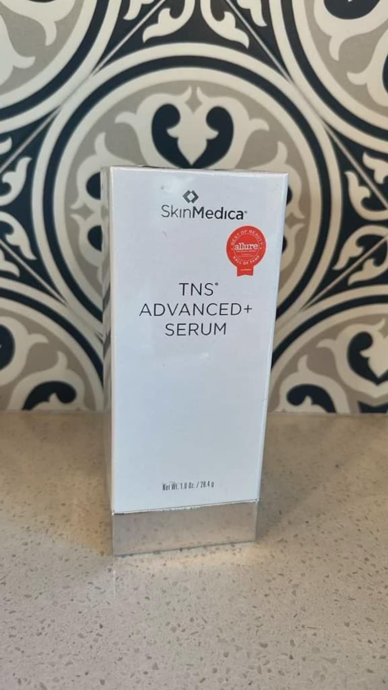 May include: A white rectangular SkinMedica TNS Advanced+ Serum box with a silver base. The front features the brand logo, product name, and a red Allure Best of Beauty seal. The text at the bottom indicates a net weight of 1.0 oz. / 28.4 g.