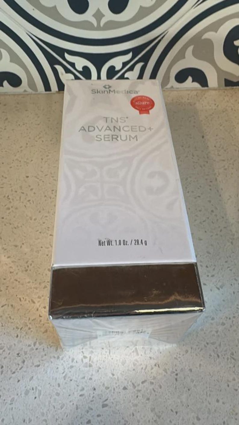 May include: A white rectangular SkinMedica TNS Advanced+ Serum box with a metallic silver base. The front features the text SkinMedica, TNS Advanced+ Serum, and a red Allure award seal. The net weight is printed as 1.0 oz. / 28.4 g on the front.