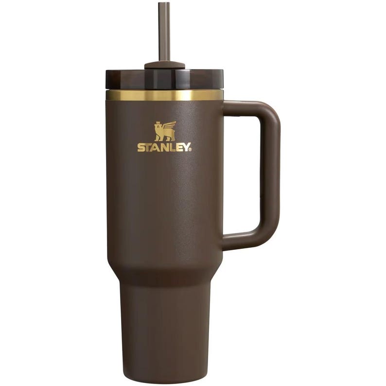 May include: A dark brown Stanley tumbler with a matching handle, a gold-toned logo featuring a winged bear, a gold-colored rim, a dark translucent lid, and a central straw. The cylindrical vessel has a tapered base and a sturdy side handle.