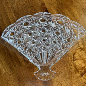May include: A clear pressed glass dish shaped like an open hand fan. The surface features a geometric pattern of starbursts and circular cutouts, with a ribbed shell-like handle at the base that includes the embossed text "NRS" in a script font.