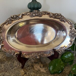 May include: An oval silver-toned serving platter featuring a polished, reflective center basin and a wide, ornate border decorated with raised floral motifs and scrolling filigree patterns along the entire outer rim of the metal tray.