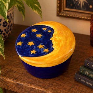 Puede incluir: Un tarro redondo de cerámica con tapa que presenta un diseño pintado a mano de una luna creciente amarilla sobre un cielo nocturno azul profundo lleno de estrellas amarillas. La tapa es abovedada y la base es de un color azul oscuro sólido.