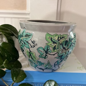May include: A round ceramic planter with a glossy light gray finish, featuring a hand-painted design of green and teal ivy leaves with dark outlines. The pot has a wide, circular opening and a smooth, curved body, showcasing a botanical motif.