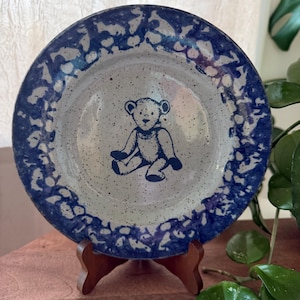 May include: A round ceramic plate featuring a speckled light gray center with a dark blue illustration of a seated teddy bear. The rim of the plate is decorated with a wide, dark blue sponge-painted pattern that contrasts with the speckled base.