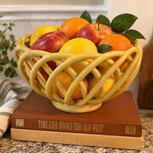 Puede incluir: Un cuenco de cerámica amarilla con un diseño de celosía trenzada lleno de fruta fresca, incluyendo manzanas rojas, naranjas y un limón. El cuenco descansa sobre dos libros de tapa dura apilados titulados Time-Life Books The Old West y A Country Home.