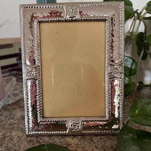 May include: A rectangular silver-toned picture frame featuring a hammered metal texture, rope-style inner borders, and decorative embossed floral accents at the centre of each side. The frame surrounds a plain, light beige rectangular insert.