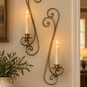 May include: Two wall-mounted candle holders made of antique brass-toned metal. Each holder features a long, curved, scroll-like design with a circular base for a single taper candle. Both holders are currently holding lit white taper candles.