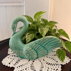 May include: A glossy turquoise ceramic swan planter featuring a long, curved neck and detailed wing feathers. The swan is filled with a trailing pothos plant that has variegated green and white heart-shaped leaves, creating a vibrant contrast.