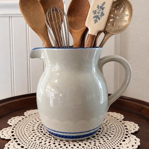 May include: A light grey ceramic pitcher with a blue rim and a white lace-like pattern near the base, filled with various kitchen utensils including wooden spoons, a metal whisk, a spatula with a blue floral design, and a slotted metal spoon.