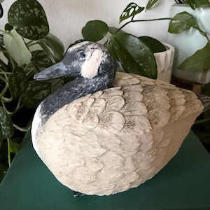 Op de afbeelding: Een decoratief ganzenbeeldje met een zwarte kop en nek en een witte wangvlek. Het lichaam is getextureerd met uitgesneden veertjes in een lichtbeige kleur. De vogel is afgebeeld in een rustende houding met de kop opzij gedraaid.