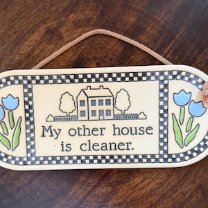 May include: An oval ceramic wall plaque with a cream background, featuring a black and white house illustration, blue tulips, and a black checkered border. The text reads, My other house is cleaner. It includes a tan cord for hanging purposes.