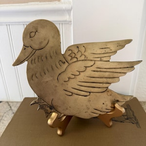 May include: A flat, brass-toned metal duck figurine featuring etched line details on the wings and body. The duck is depicted in a side profile with its wings slightly spread and is supported by a small, gold-colored, three-legged display stand.